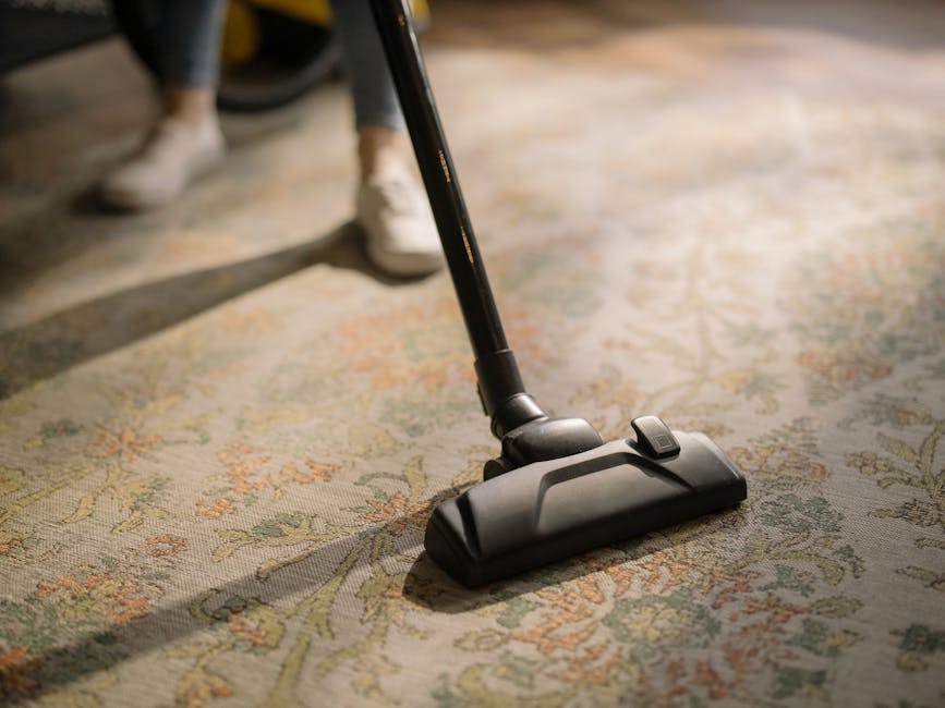 A person kneeling on a patterned carpet in a home environment, using a yellow and black vacuum cleaner with a transparent dust container to perform surface cleaning and deep cleaning. Their hands are 