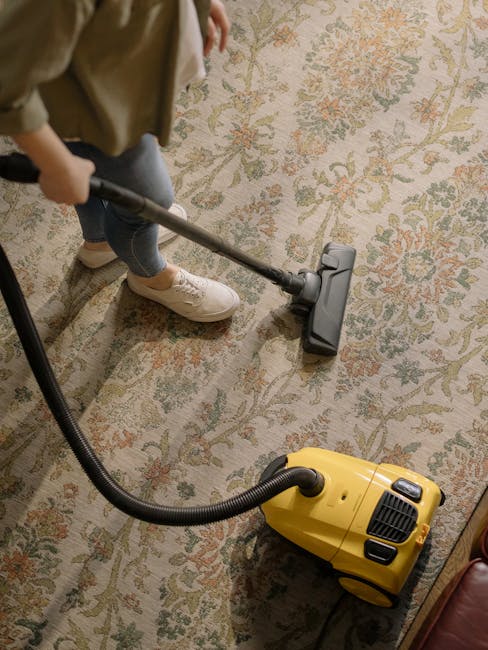 A child using a hand-held vacuum cleaner to clean an ornate, patterned area rug with floral motifs in a domestic living room setting. The carpet features a mix of beige, green, and orange tones, and a