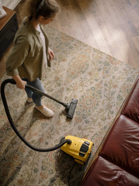 A child using a hand-held vacuum cleaner to clean an ornate, patterned area rug with floral motifs in a domestic living room setting. The carpet features a mix of beige, green, and orange tones, and a
